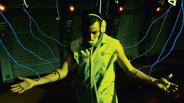 Still from the film Sleep Dealer (2008) showing a man in a face mask with his arms spread wide and connected to glowing tubes