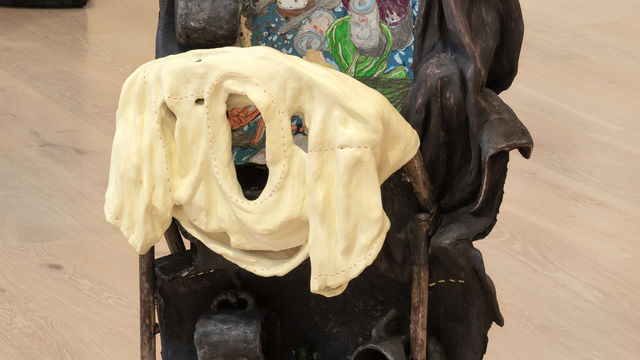 A sculpture with an combination of an upright steel folding chair encased within a tower of five tires  cut in half.  A butter yellow ceramic shirt is draped over the folded steel chair.