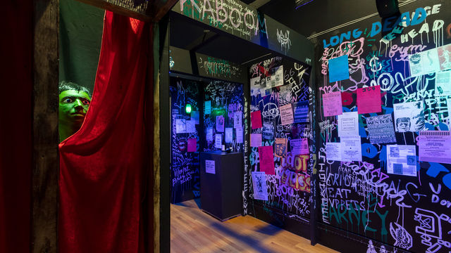 A green head pokes out from behind a red curtain in a flyer- and graffiti-covered haunted house