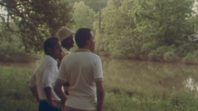 Rectangular image of three black men standing on a grassy, green riverbank. The brownish river splits the image in three, with the other side of the riverbank filled with trees. The men stand in a line, with the middle and far right men fully visible and the head of the far left man visible. That man has a cream white hat on. The two men we can see have their arms on their hips. We cannot see their legs.